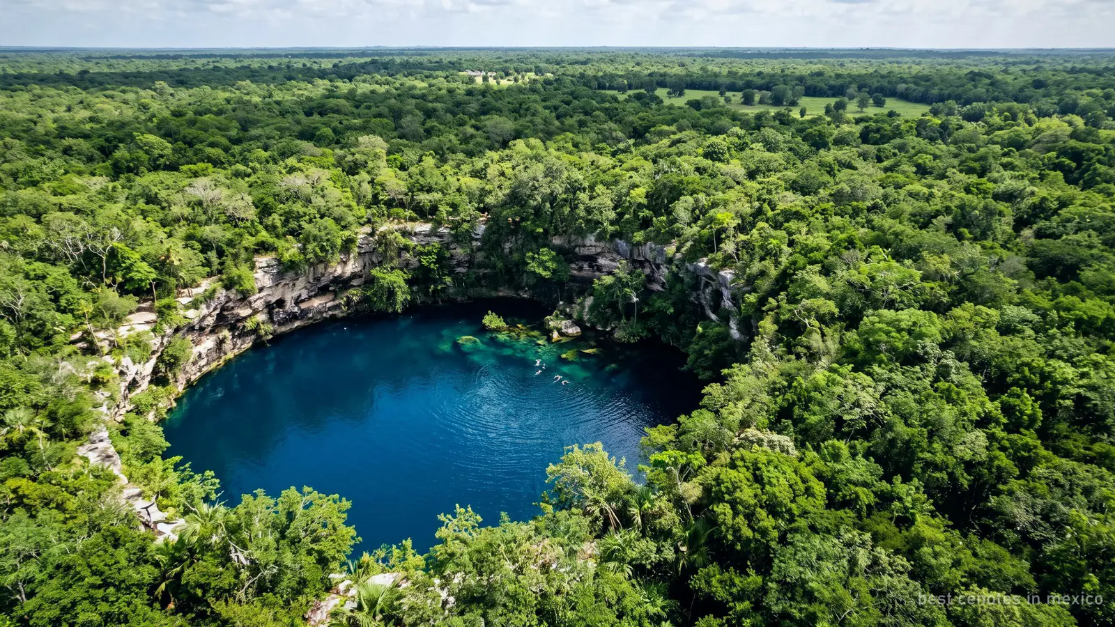 Aerial view of a round open cenote, deep blue water ringed by limestone and jungle